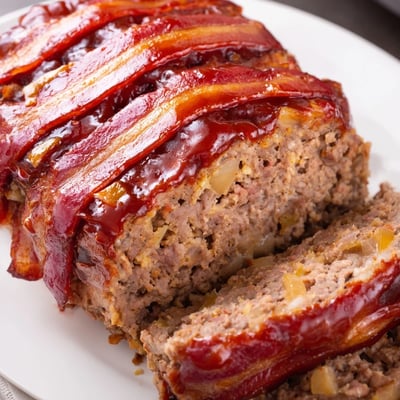 Freshly baked BBQ Beef Bacon Wrapped Meatloaf resting on a wooden board, ready to serve with roasted vegetables.